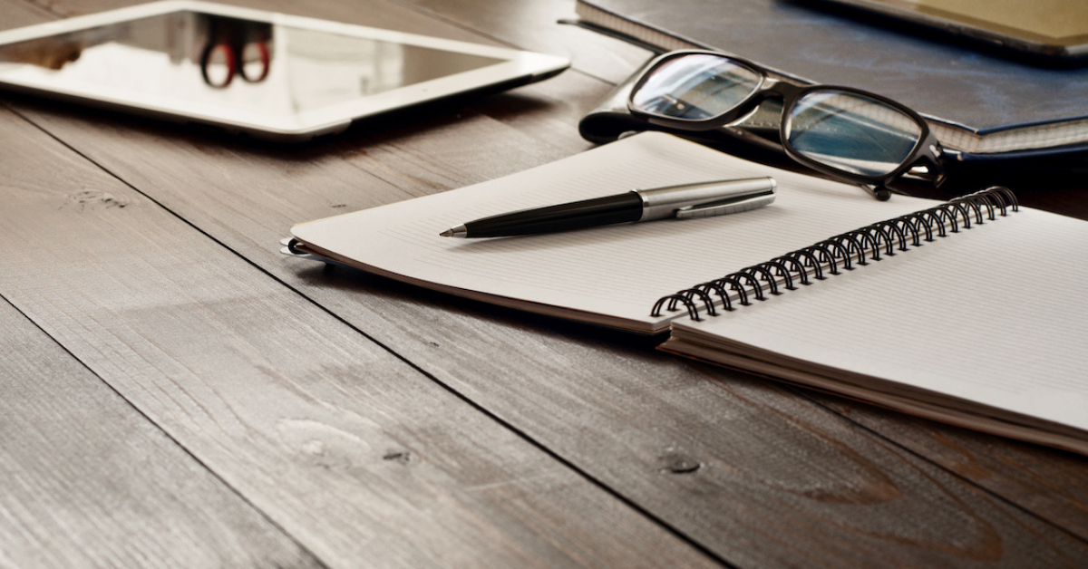 wooden desk with ipad, notebook, a pen, and a pair of glasses on top of desk